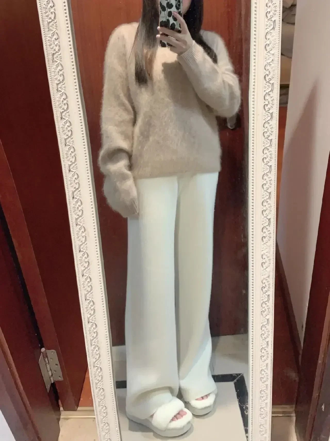 Kvinde i spejl iført beige sweater og hvide, brede bukser. Moderne outfit, minimalistisk stil, komfortabelt tøj, mode til efterår/vinter.