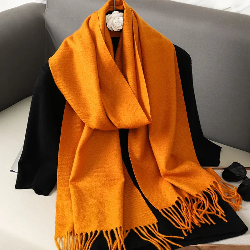 Orange uldtørklæde med frynser draperet over sort jakke på grå sofa. Stilfuldt vintertilbehør, varm og elegant. Perfekt til kolde dage.