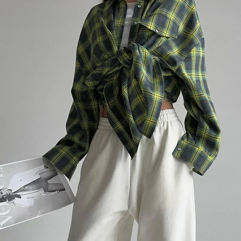 Kvinde iført grønternet skjorte bundet foran og hvide bukser, holder et modeblad. Moderne streetwear-stil, trendy tøj, afslappet look.