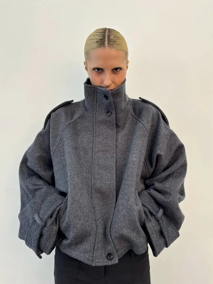 Kvindelig model iført grå oversized jakke med høj krave og sorte knapper, minimalistisk mode, moderne stil, hvid baggrund.