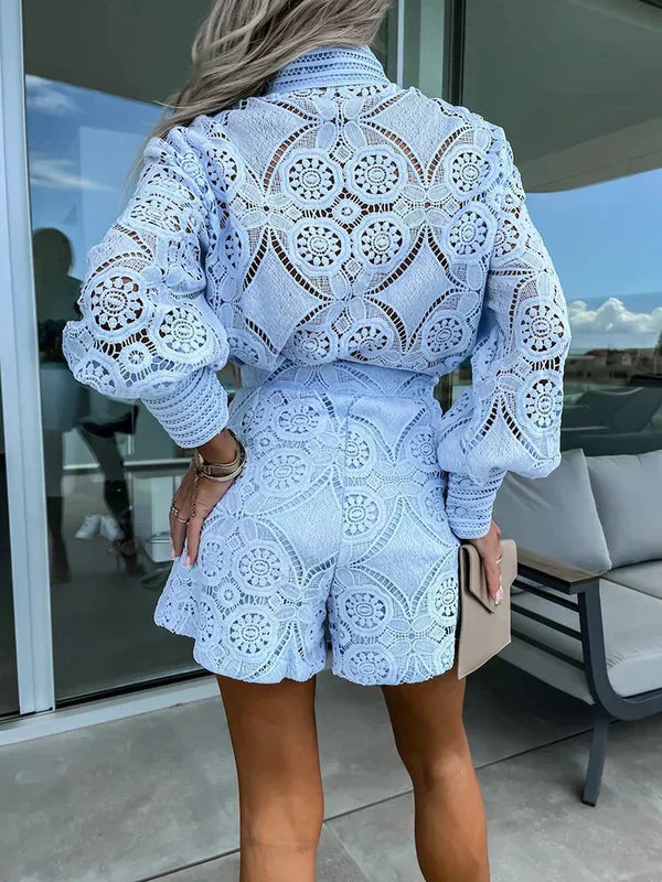 Kvinde i lyseblåt, hæklet sæt med lange ærmer og shorts, står på terrasse med glasvæg. Moderne sommermode, elegant design, udendørs baggrund.
