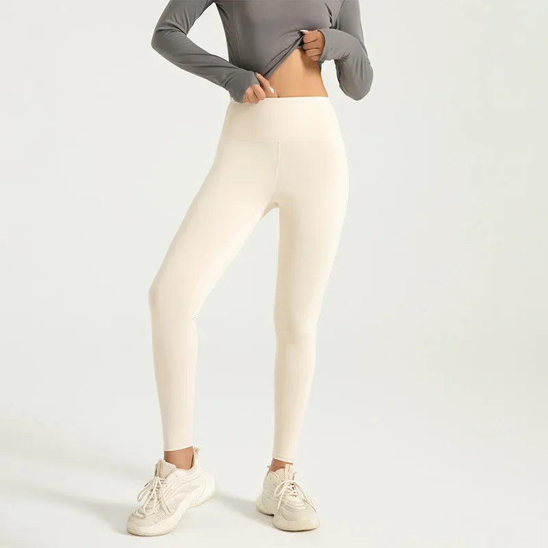 Kvinde iført beige leggings og grå langærmet top, poserer mod hvid baggrund. Komfortabelt sportstøj, ideelt til yoga og fitness.
