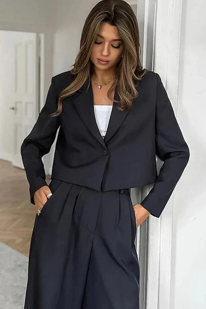 Kvinde iført sort jakkesæt med hvid top, står i moderne interiør. Stilfuldt business-look, mode, elegant tøj, professionel påklædning.