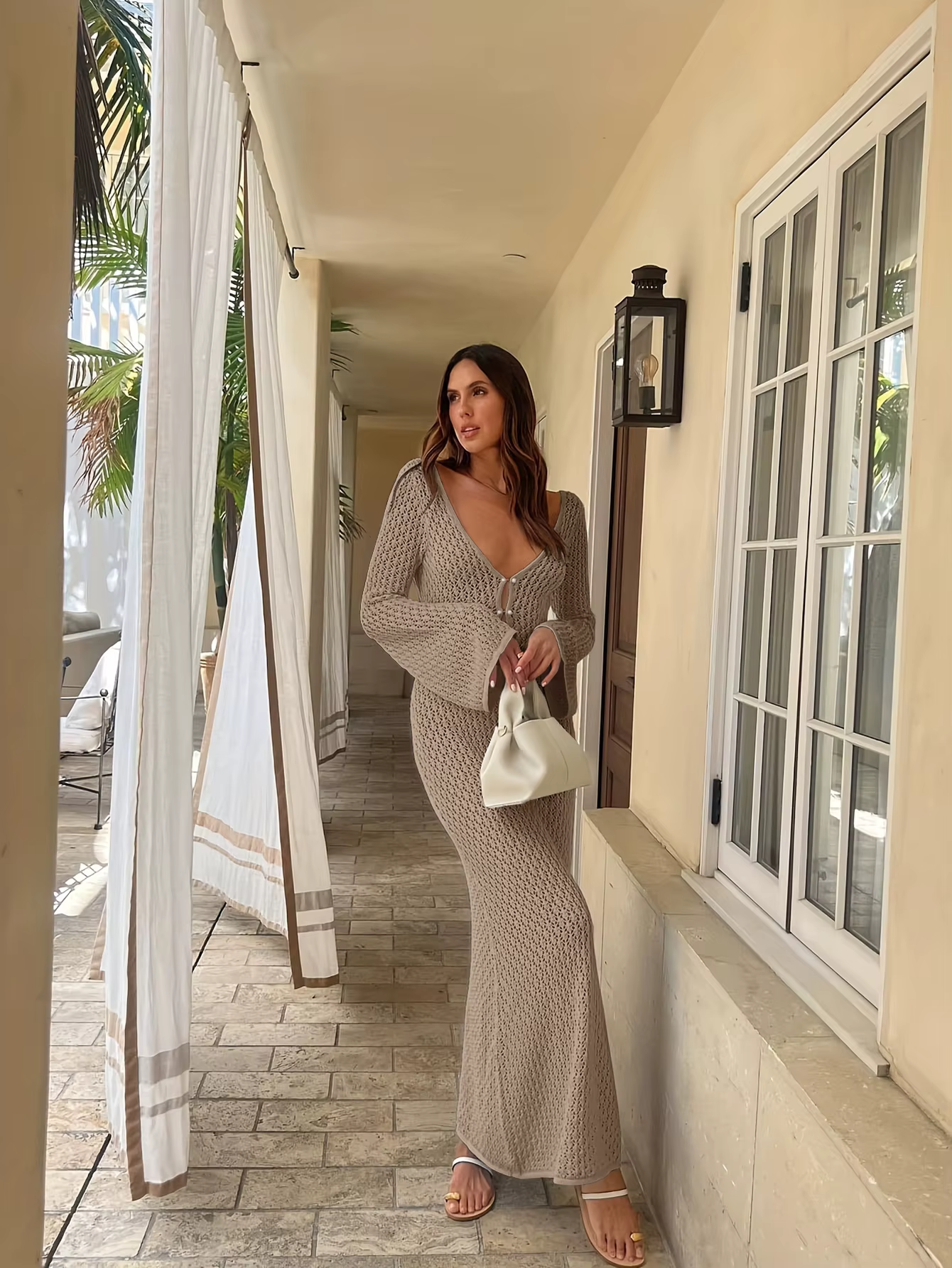 Kvinde i lang beige strikkjole med dyb udskæring, holder hvid taske, står på terrasse med søjler og gardiner, tropisk baggrund.