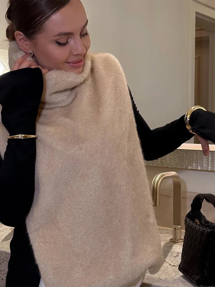 Kvinde iført beige strikponcho og sort trøje, smilende ved køkkenvask. Elegant mode, vintertøj, moderne stil, gyldne armbånd.