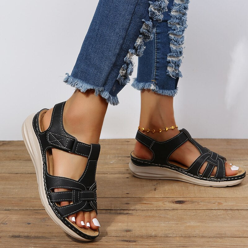Kvinde iført sorte lædersandaler med velcrolukning og komfortabel sål, kombineret med blå jeans med frynser. Perfekt til sommermode og afslappet stil.