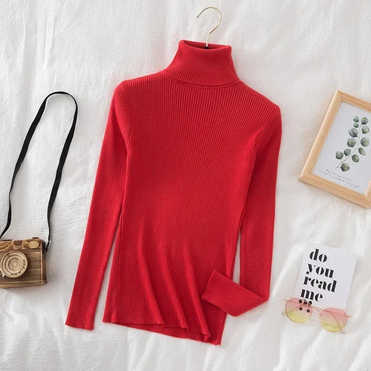 Rød rullekrave sweater på hvidt sengetøj, ved siden af retro kamera taske, solbriller og en ramme med eukalyptusblad. Mode, efterår, stil.