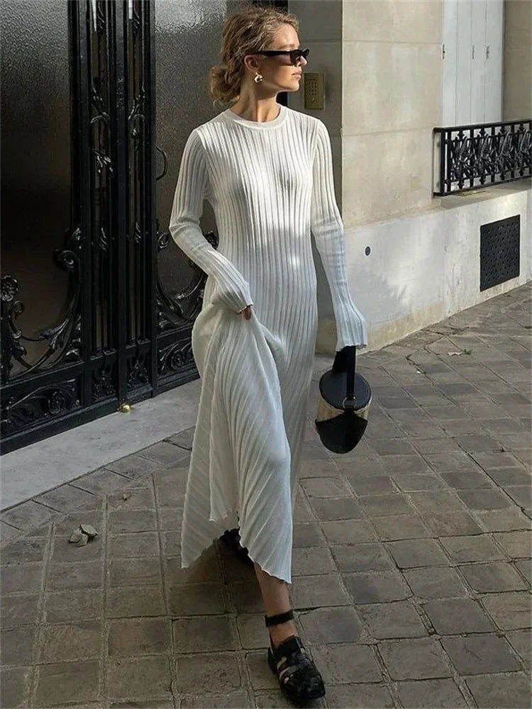 Kvinde i hvid, ribstrikket kjole går på brostensbelagt gade, bærer sort taske og solbriller. Elegant mode, moderne stil, byliv.