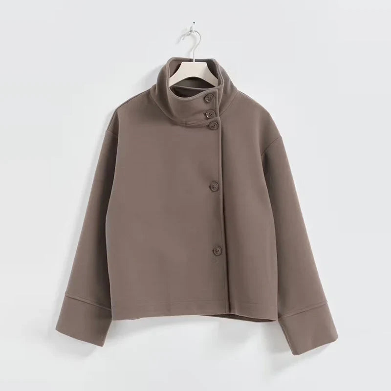 Brun jakke med asymmetrisk knaplukning og høj krave, hængende på bøjle. Moderne design, minimalistisk stil, perfekt til efterårsmode.