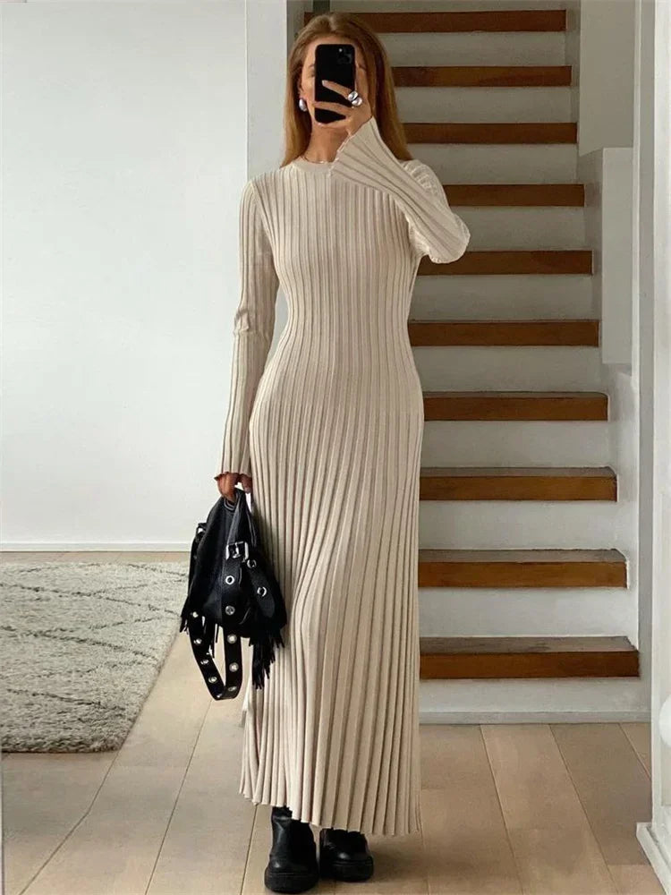 Langærmet beige ribstrikket kjole, kvinde står foran trappe, sort taske i hånden, moderne minimalistisk stil, trendy mode, indendørs.