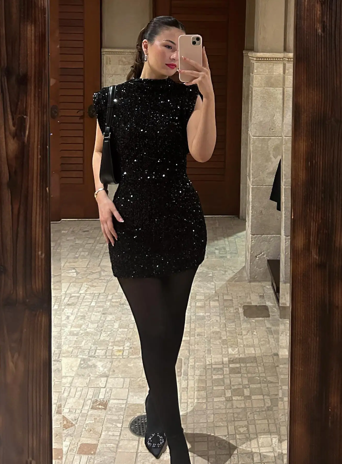Kvinde i sort glitrende kjole tager selfie i spejl. Elegant aftenlook med sort taske og strømpebukser. Perfekt til fest eller gallaaften.