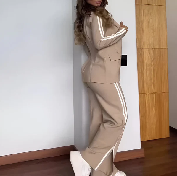 Kvinde iført beige jakkesæt med hvide striber, poserer ved væg. Moderne mode, stilfuldt design, komfortabelt tøj, trendy look.