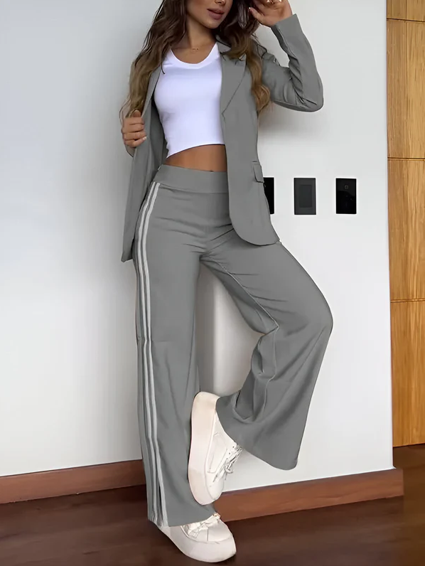 Kvinde iført grå jakkesæt med hvide striber, hvid top og hvide sneakers, står op ad en hvid væg. Moderne damebeklædning, casual business look.