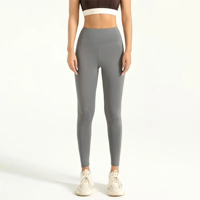 Kvinde iført grå højtaljede leggings og hvid sportstop, stående mod lys baggrund. Komfortabelt træningstøj, perfekt til fitness og yoga.