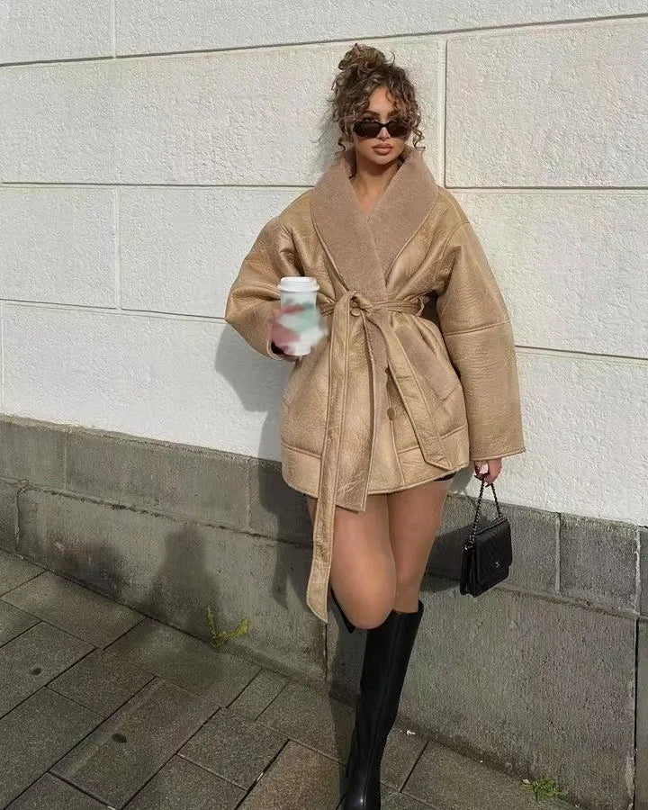 Kvinde i beige frakke, sorte solbriller og støvler, holder en kaffekop og en sort taske, står mod en hvid mur. Mode, efterår, street style.