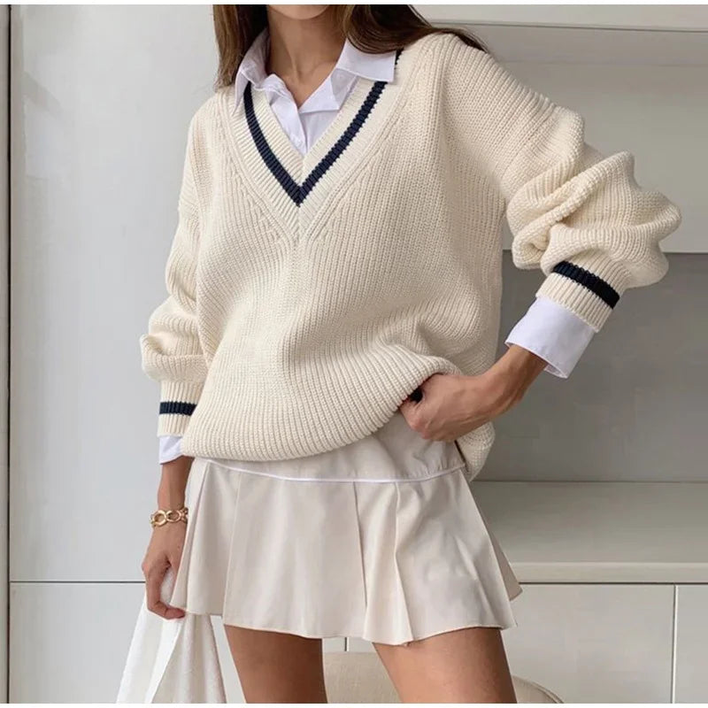 Kvinde iført hvid striktrøje med V-hals og blå striber, kombineret med hvid skjorte og kort plisseret nederdel. Mode, efterårstøj, stilfuldt look.