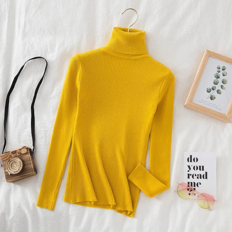 Gul rullekrave sweater på hvidt sengetøj, ved siden af retro kamera, solbriller og en bog. Moderne mode, efterårstøj, stilfuldt design.