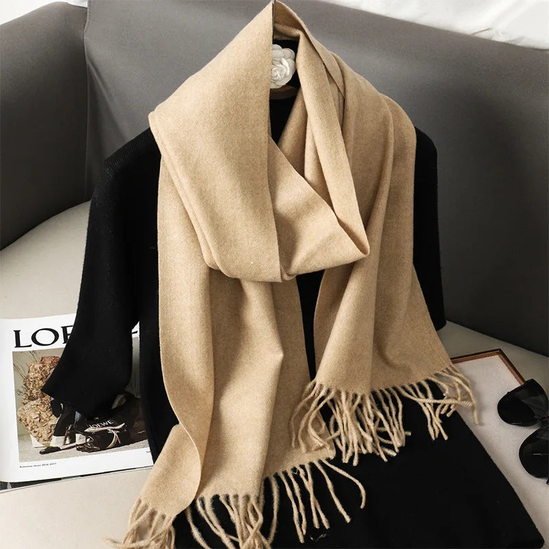 Beige uldtørklæde med frynser draperet over sort sweater på sofa. Elegant og stilfuldt tilbehør til vintermode.