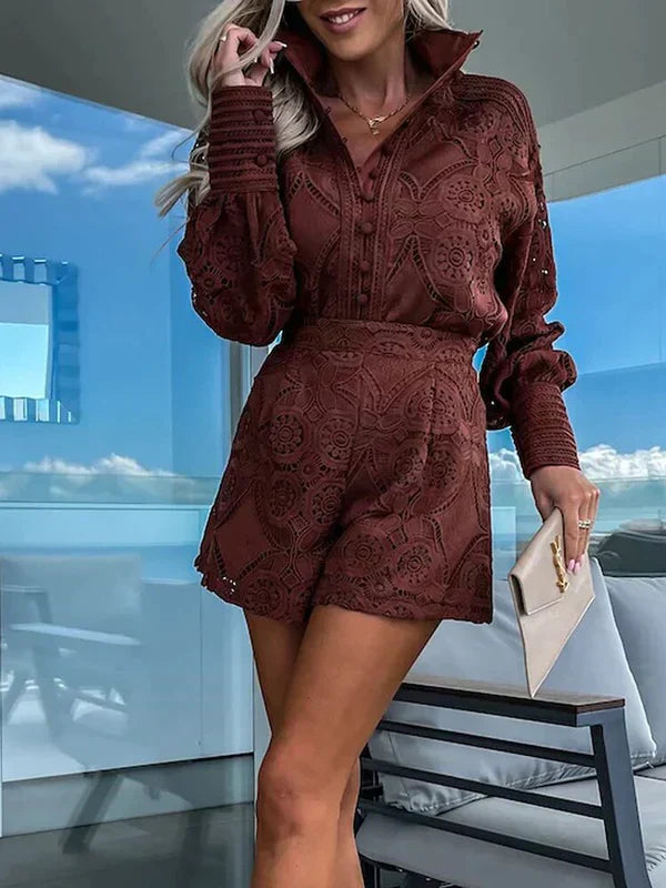 Kvinde iført brun, mønstret jumpsuit med lange ærmer, står på altan med blå himmel i baggrunden. Holder beige clutch. Mode, sommer, stil.