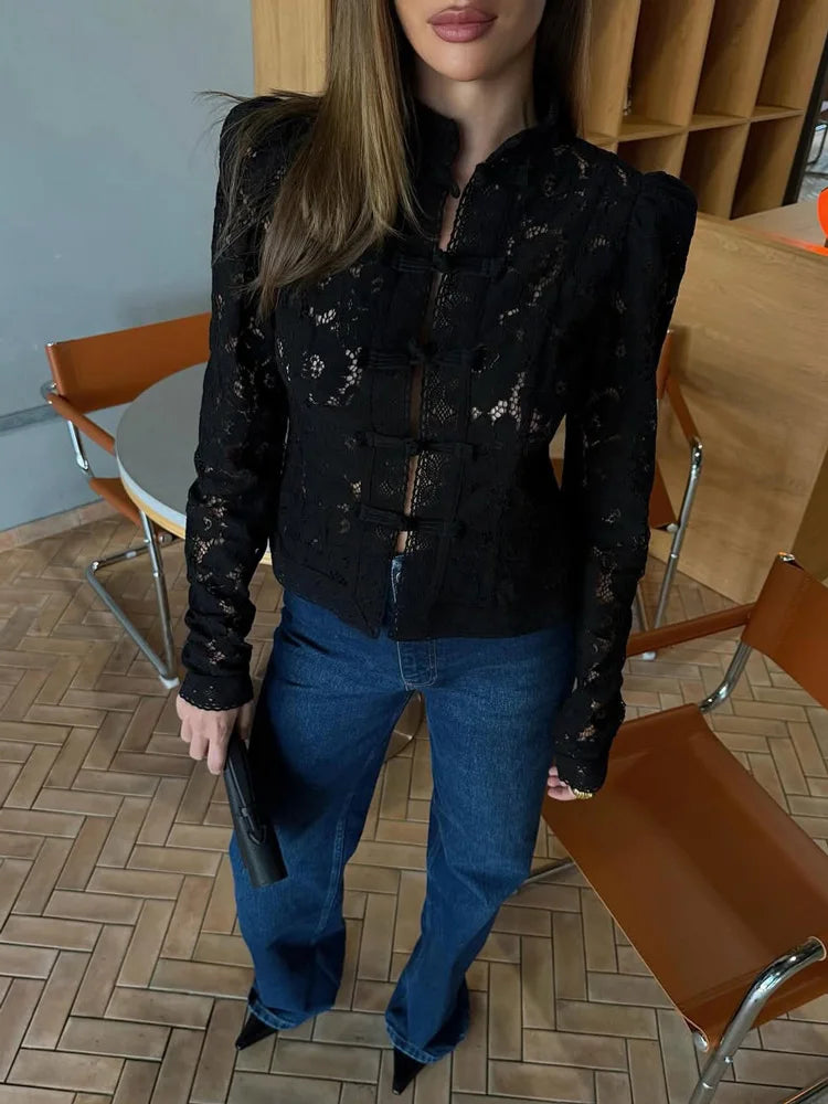 Kvinde iført sort blondejakke og blå jeans, står ved et bord med brune stole. Mode, stilfuldt tøj, moderne kvindemode, casual outfit.