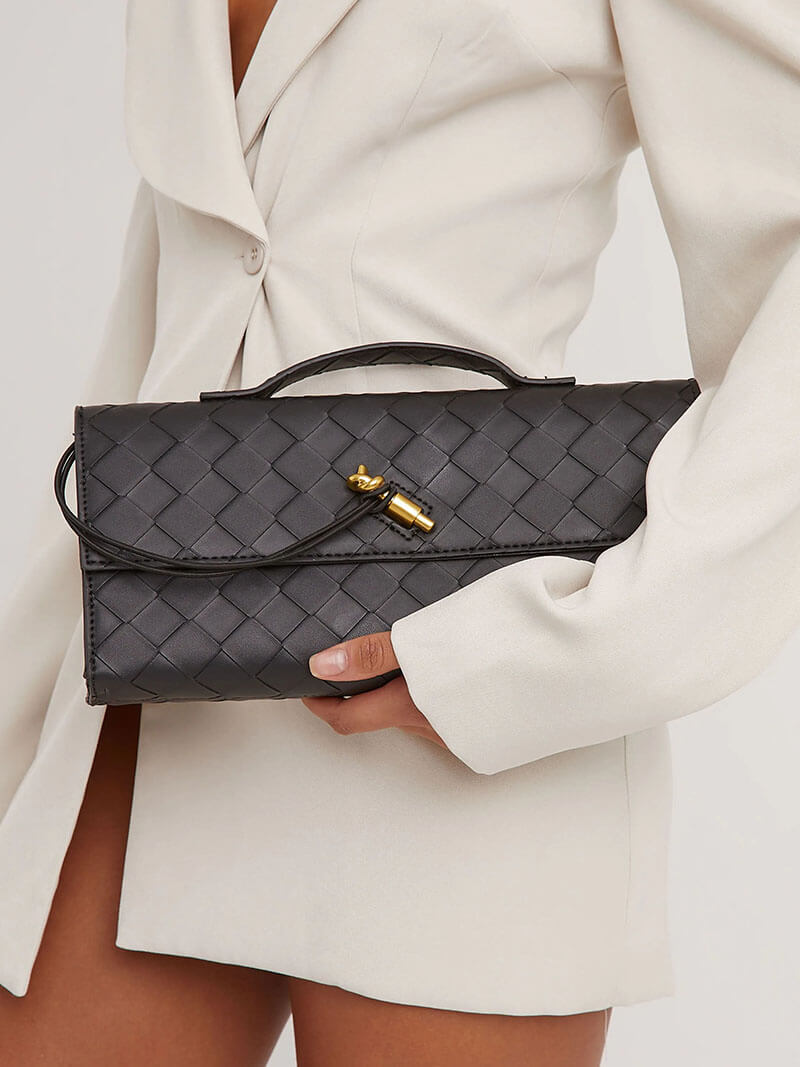 Kvinde i hvid blazer holder sort flettet clutch-taske med gulddetaljer. Elegant modeaccessory, perfekt til stilfulde begivenheder.