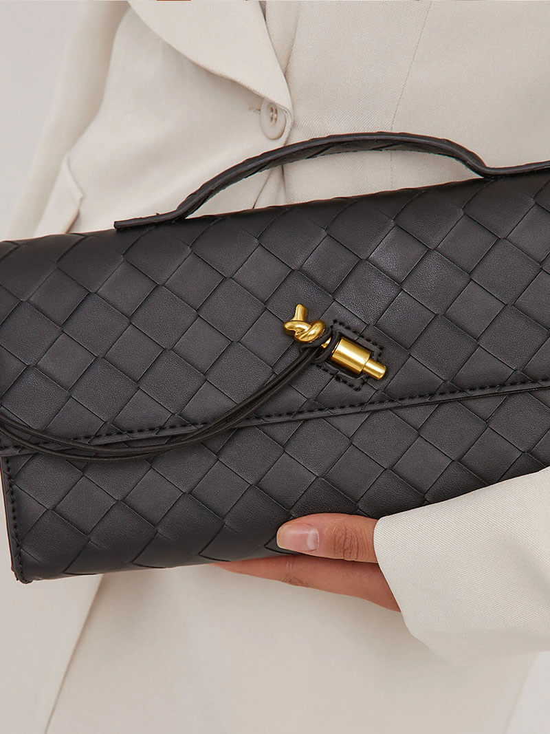 Sort flettet læder clutch med guldspænde, holdt af person i hvid jakke. Elegant håndtaske, perfekt til aftenbrug og modebevidste kvinder.