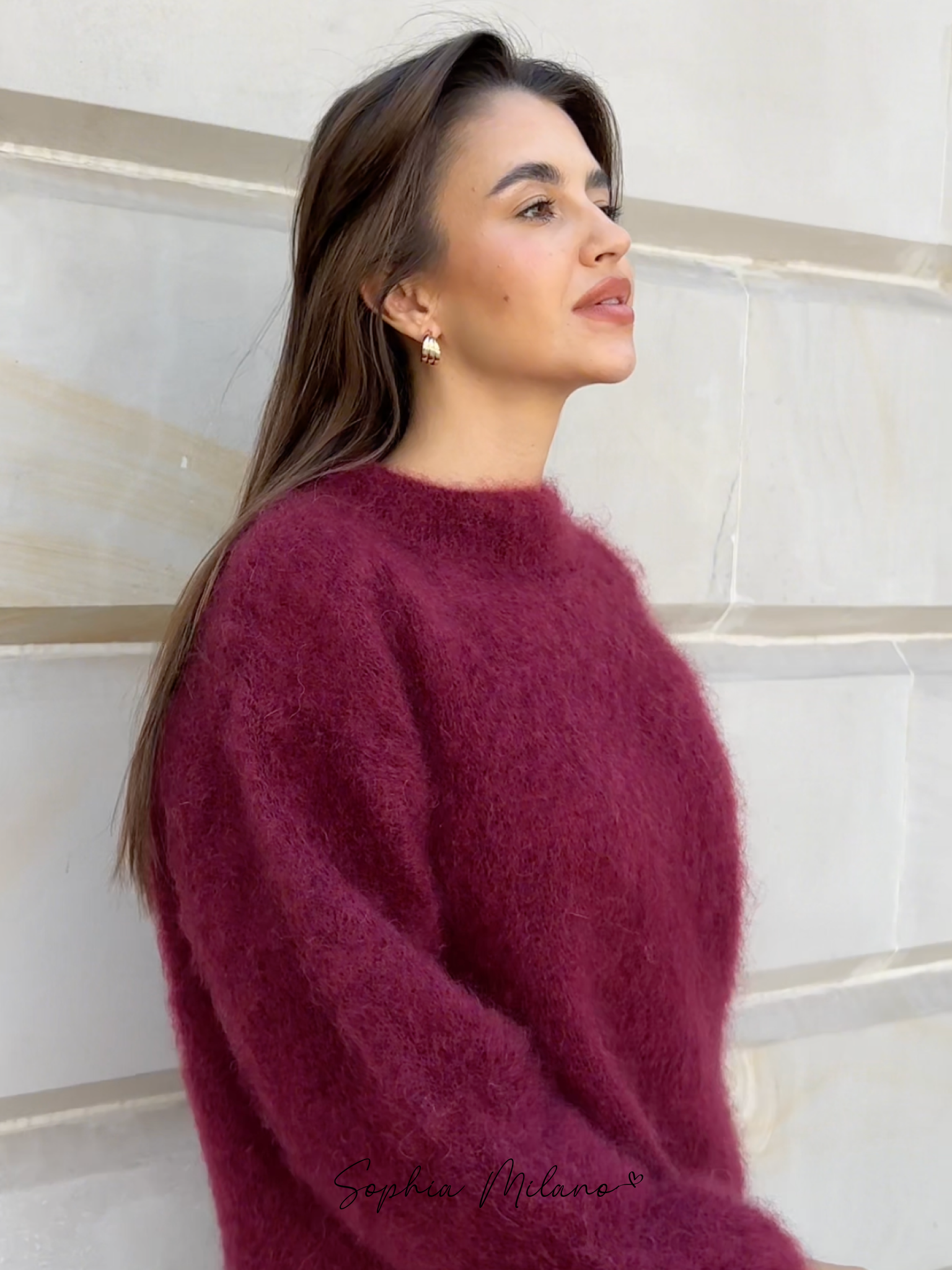 Kvinde iført bordeaux striktrøje, poserer mod lys murstensvæg. Elegant efterårslook, mode, stilfuldt outfit, moderne kvindetøj.