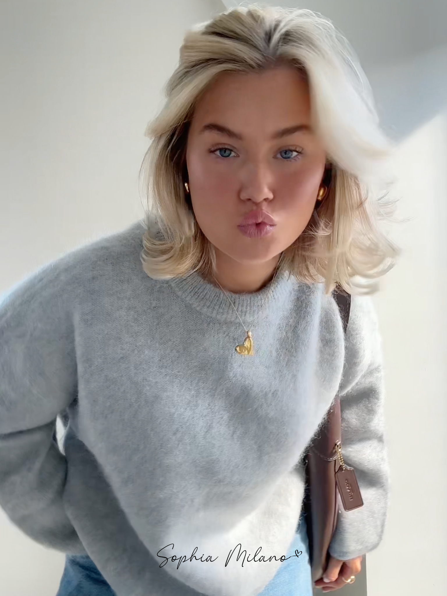 Jeg ved ikke, hvem personen er, men billedet viser en kvinde i en grå sweater med blond hår, der poserer indendørs. Hun bærer en halskæde med et hjertevedhæng.