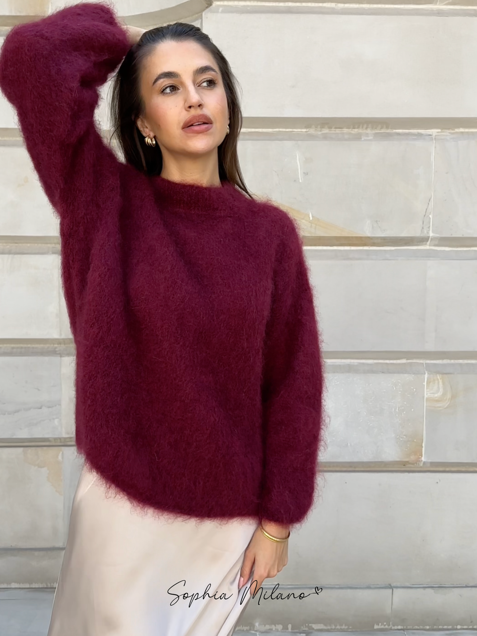 Kvinde iført bordeaux uldtrøje og hvid nederdel foran beige væg. Elegant mode, efterårslook, stilfuldt tøj, moderne kvindemode.