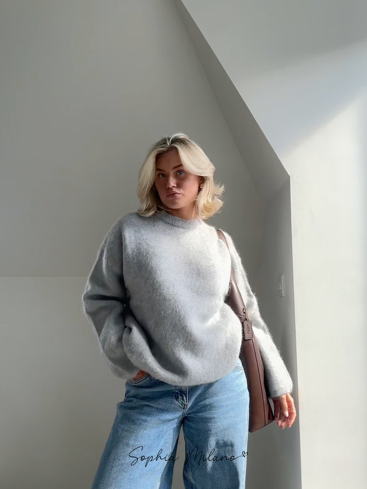 Ung kvinde i grå sweater og blå jeans står i sollys ved hvid væg. Moderne mode, afslappet stil, minimalistisk indretning.