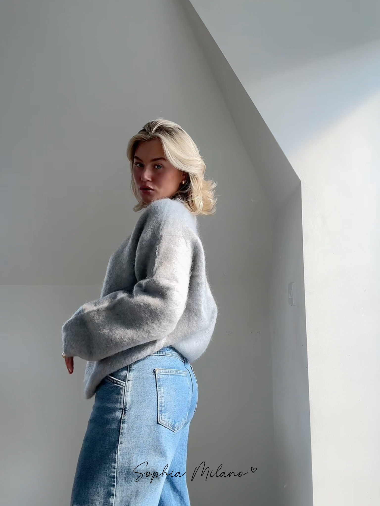 Jeg ved ikke, hvem personen er, men billedet viser en kvinde i en grå sweater og blå jeans, stående i et lyst rum med minimalistisk indretning.