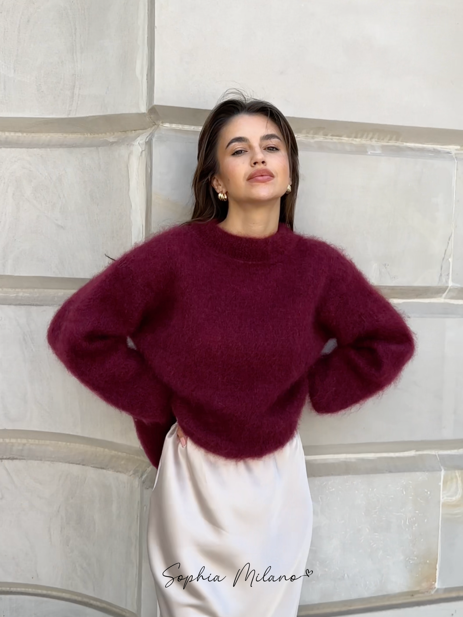 Kvinde iført bordeaux striktrøje og hvid nederdel, læner sig mod en murstensvæg. Elegant mode, efterårslook, stilfuldt outfit.