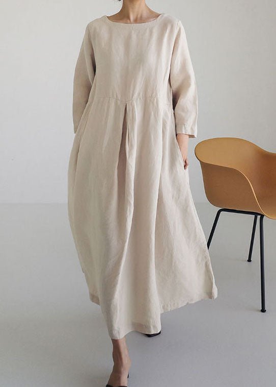 Kvinde iført lang, beige kjole med løstsiddende pasform, stående ved siden af moderne, gul stol. Minimalistisk mode, skandinavisk design.
