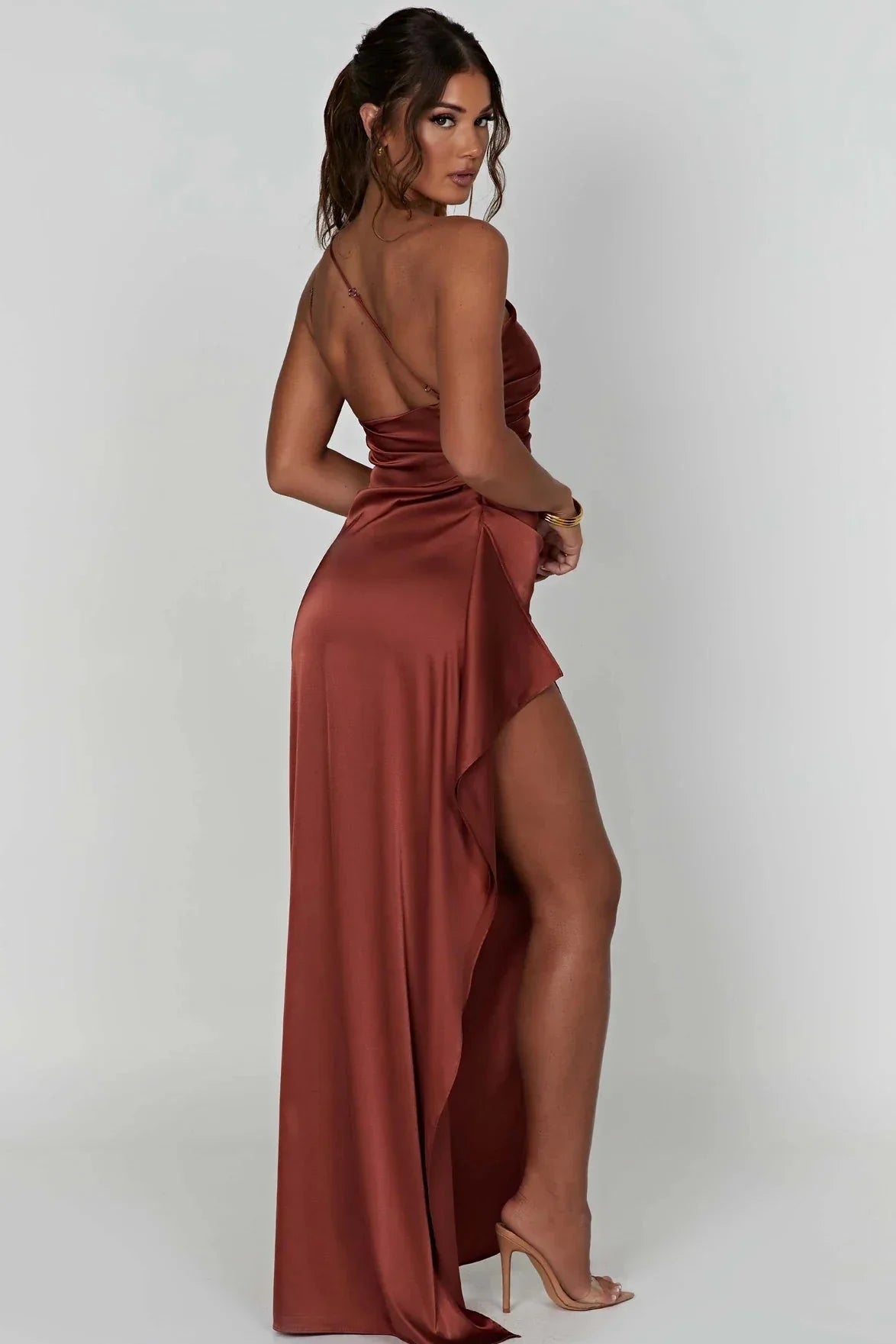 Kvinde i lang, rødbrun satin kjole med høj slids og åben ryg, poserer mod hvid baggrund. Elegant aftenkjole, mode, festkjole, stilfuldt look.