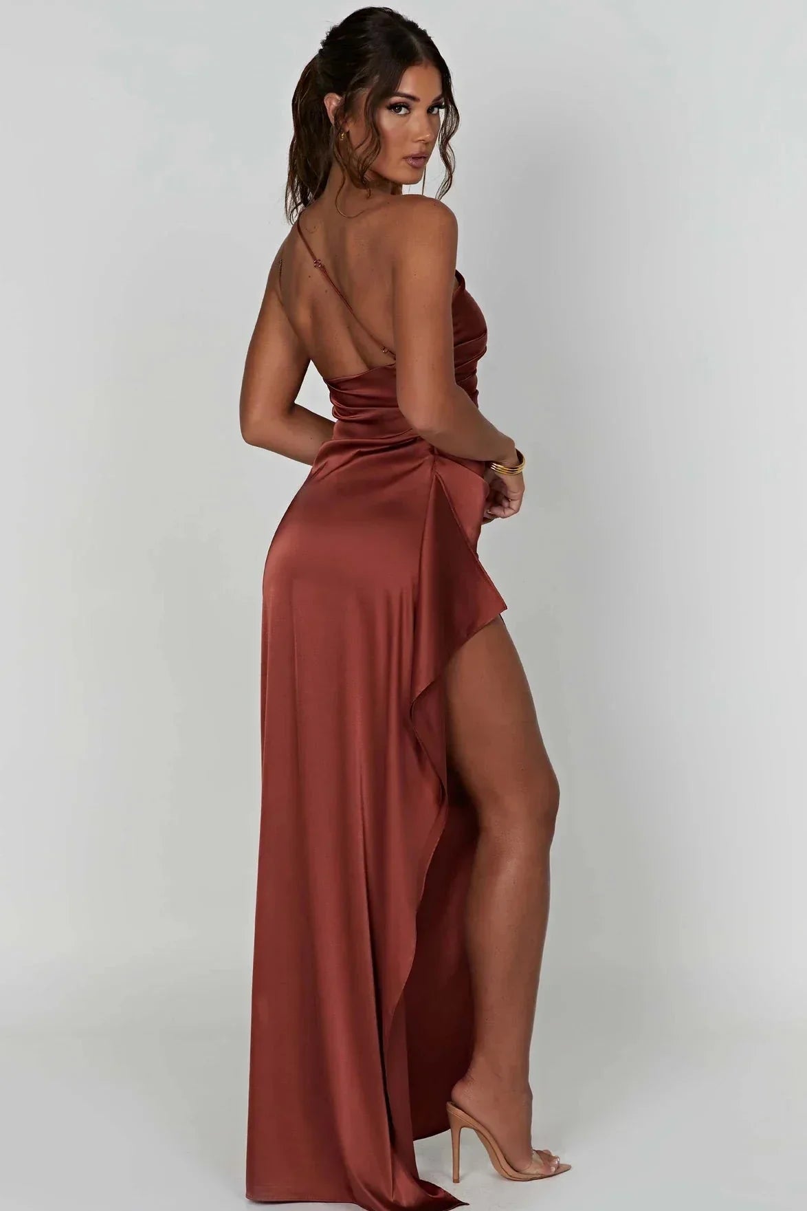 Kvinde i lang, rødbrun satin kjole med høj slids og åben ryg, poserer mod hvid baggrund. Elegant aftenkjole, mode, festkjole, stilfuldt look.
