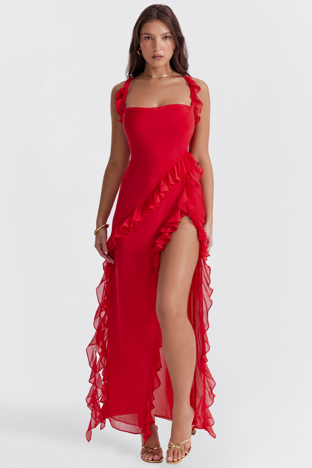 Kvinde i rød aftenkjole med flæsedetaljer og høj slids, iført guldsandaler. Elegant og stilfuldt look til festlige lejligheder.