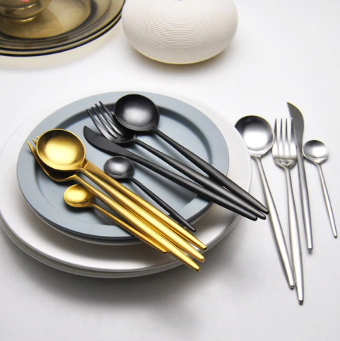 Moderne bestik i guld, sort og sølv på grå og hvide tallerkener. Elegant borddækning, stilfuldt design, rustfrit stål, minimalistisk æstetik.