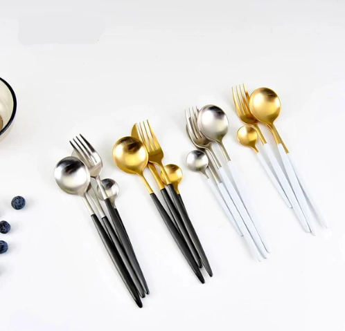 Moderne bestiksæt i guld og sølv med sorte og hvide håndtag, elegant design, skeer, gafler og knive, perfekt til stilfuld borddækning.