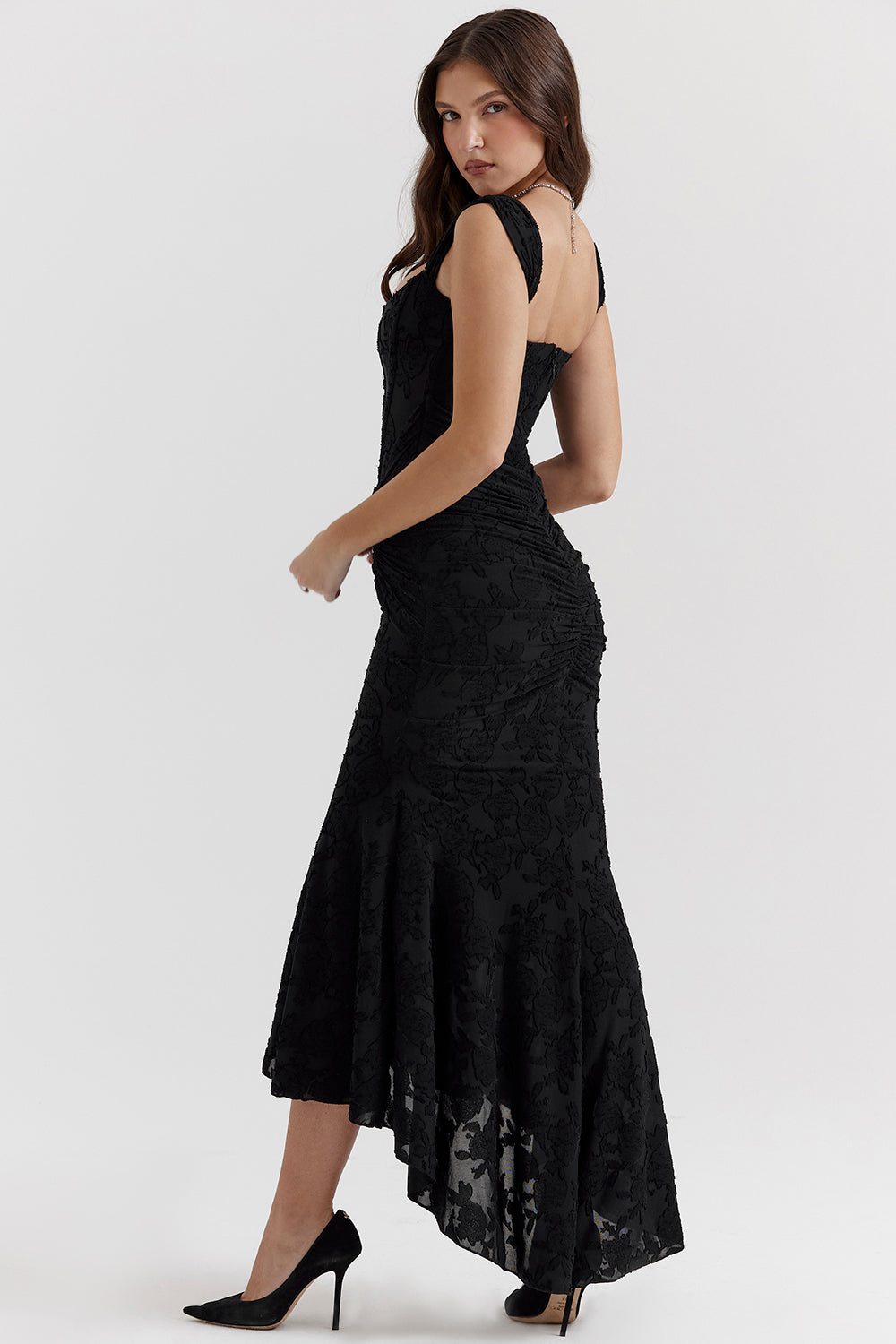 Kvinde i sort, asymmetrisk aftenkjole med blonder, poserer i profil. Elegant design, perfekt til formelle begivenheder.