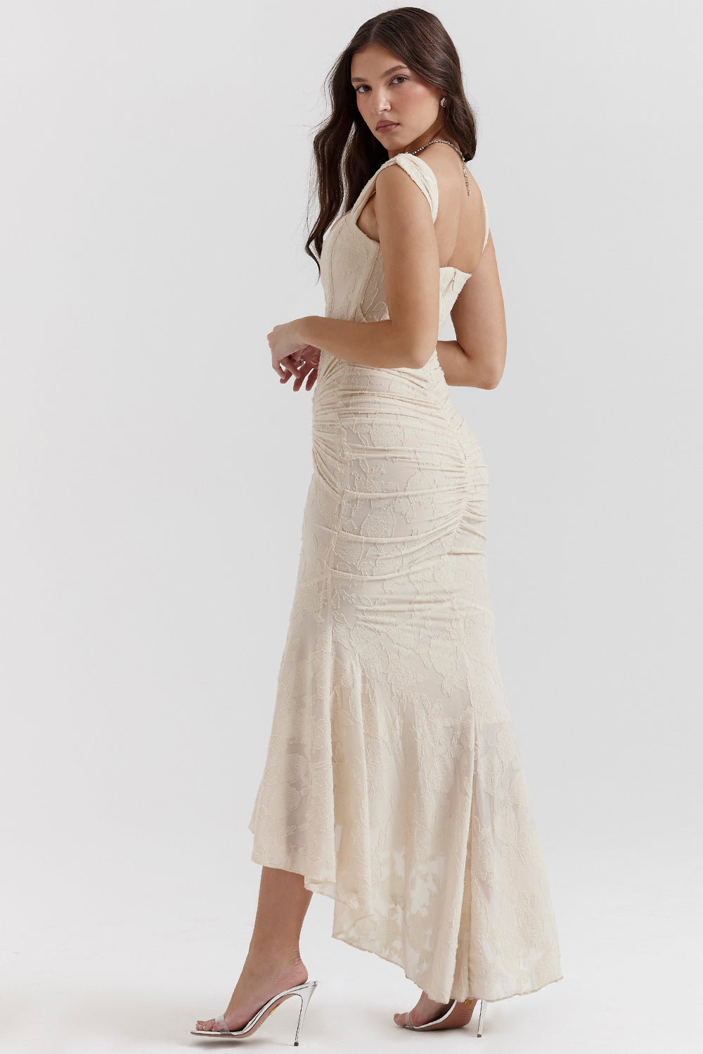 Kvinde i elegant, cremefarvet aftenkjole med asymmetrisk snit og blonder, poserer i profil. Perfekt til bryllup eller gallafest.