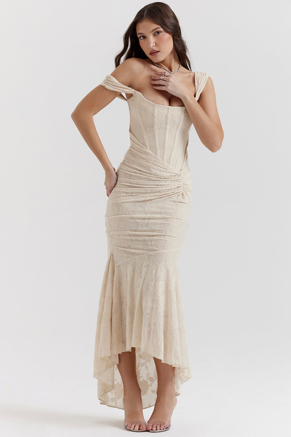 Kvinde i elegant, cremefarvet aftenkjole med asymmetrisk nederdel og off-shoulder design. Perfekt til gallafester og formelle begivenheder.