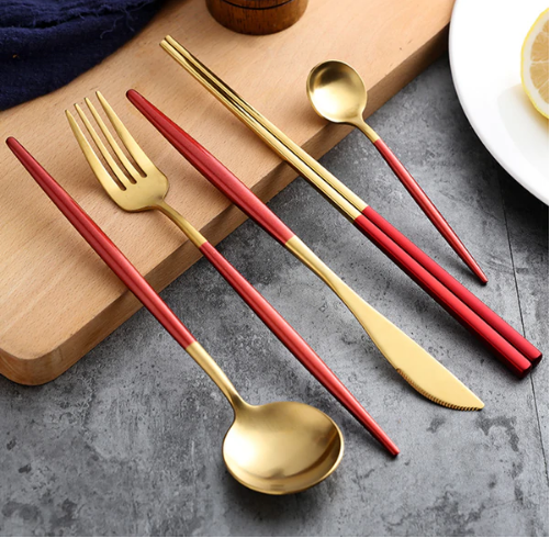 Rød og guld bestiksæt med gaffel, kniv, ske og spisepinde på grå bordplade. Elegant design, moderne køkkenudstyr, stilfuldt borddækning.
