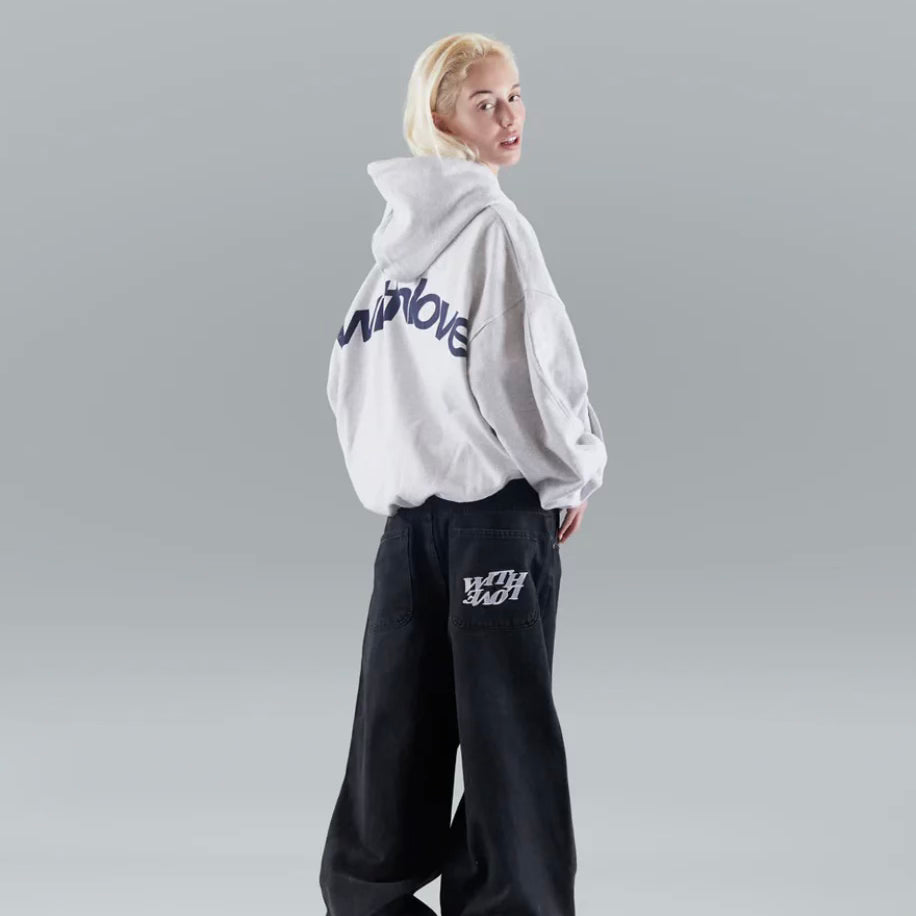 Ung kvinde i grå hættetrøje med blå skrift, poserer mod grå baggrund. Hun bærer sorte, løse bukser med hvidt tryk. Streetwear mode.
