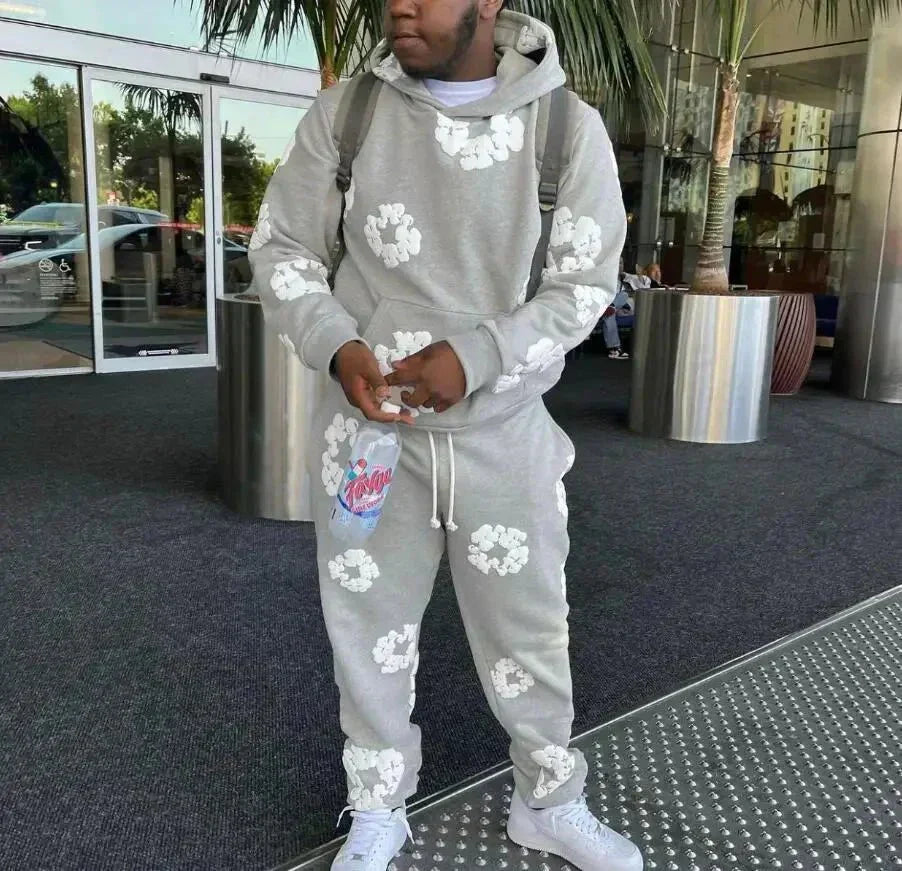 Grå hættetrøje og joggingbukser med hvide blomster, mand står foran moderne bygning. Streetwear mode, afslappet stil, urban tøj.