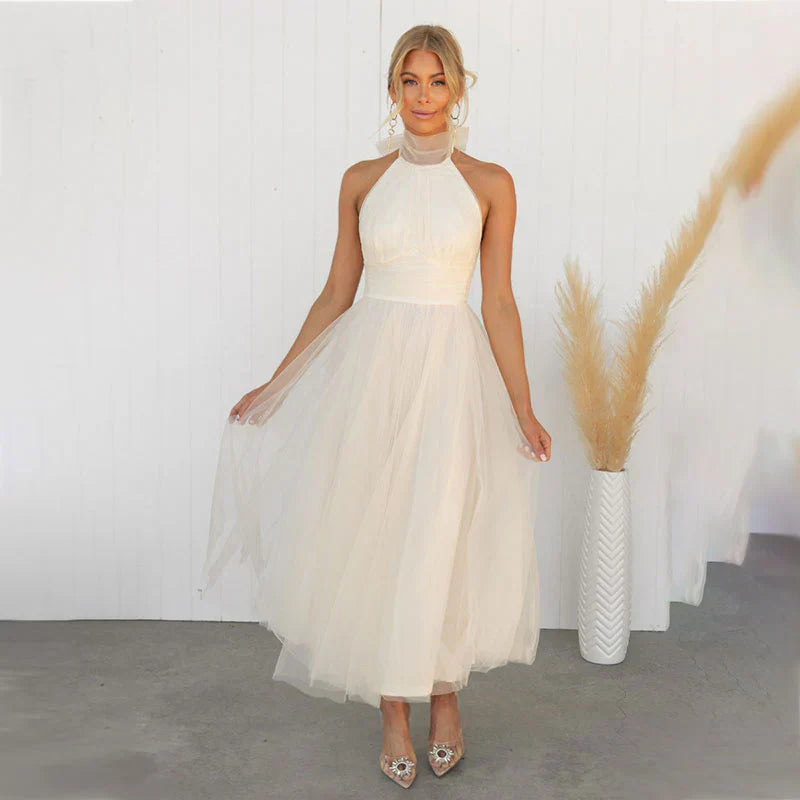 Kvinde i elegant, hvid aftenkjole med halterneck, poserer foran hvid baggrund. Tylskørt og stilfulde hæle. Perfekt til bryllup eller fest.