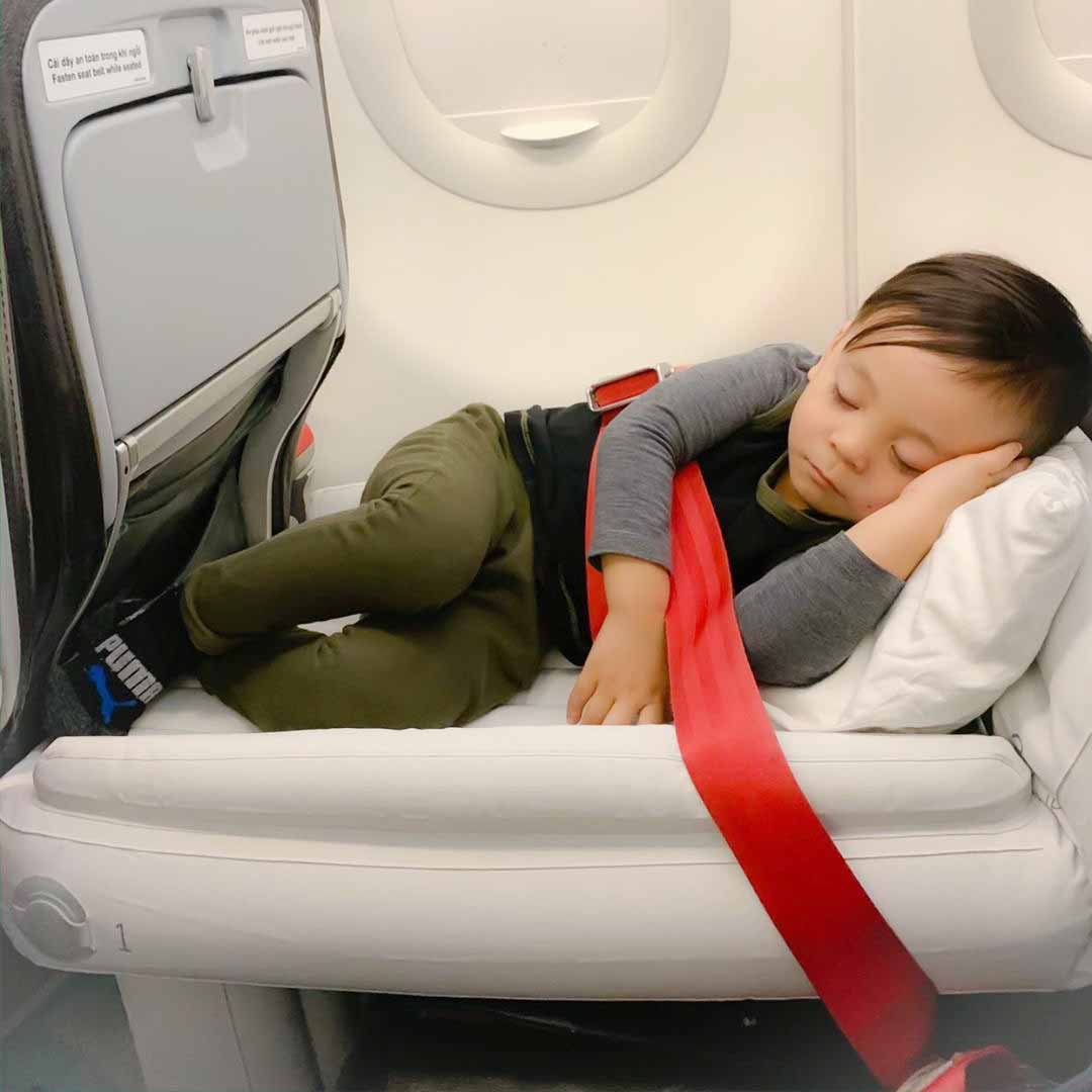 Lille barn sover fredeligt på flysæde med rød sikkerhedssele, iført grå trøje og grønne bukser. Komfortabel flyrejse, afslappet barn.