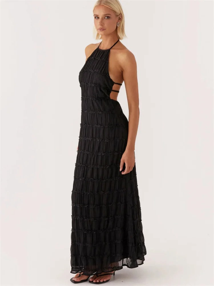 Kvinde i sort, ærmeløs maxikjole med halterneck og stropdetaljer, poserer mod hvid baggrund. Elegant aftenkjole, moderne mode.