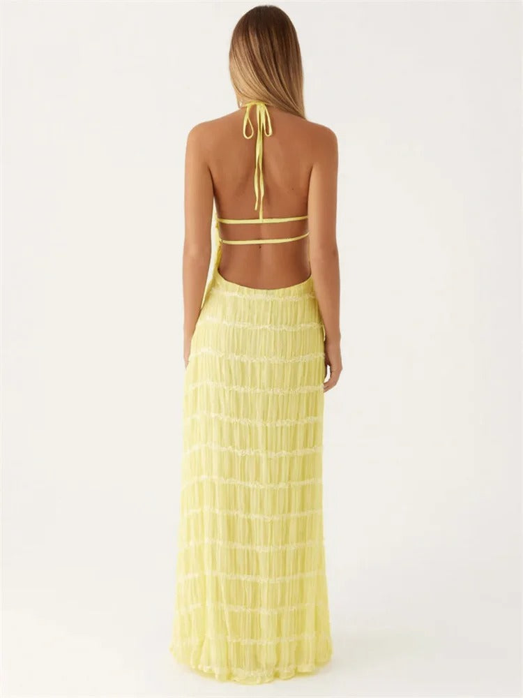 Kvinde i gul, rygfri sommerkjole med halterneck og flæsedetaljer. Perfekt til strandmode og sommerfester. Elegant og stilfuldt design.