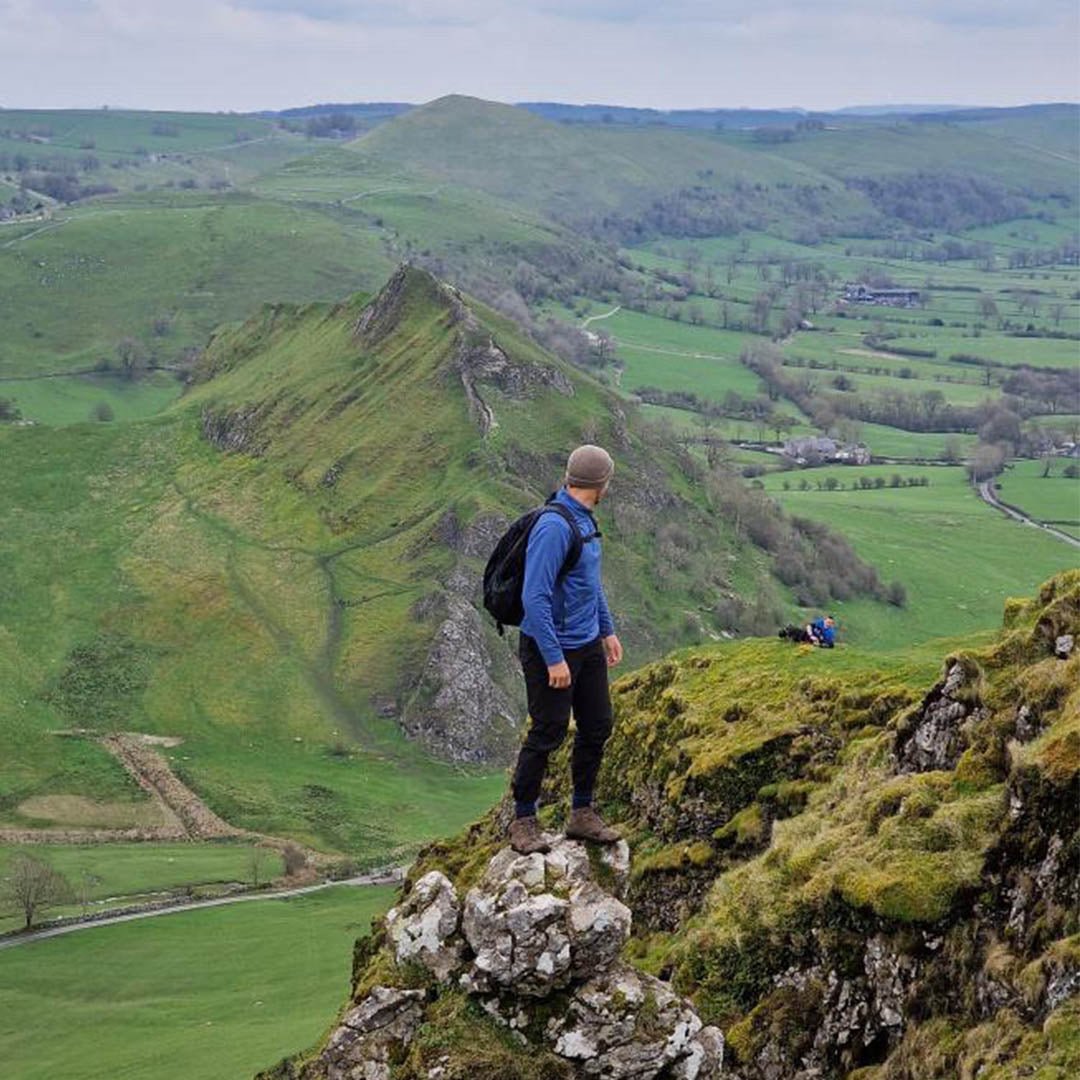 Mand på vandretur står på klippe i Peak District, England, med udsigt over grønne bakker og dale. Udendørs eventyr, natur, landskab.