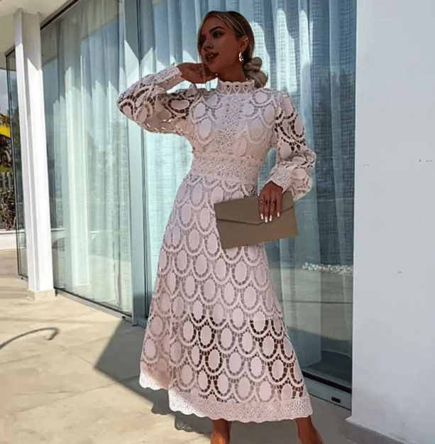 Kvindelig model i elegant, hvid blondekjole med lange ærmer, poserer udendørs ved glasvæg. Perfekt til bryllup eller fest.